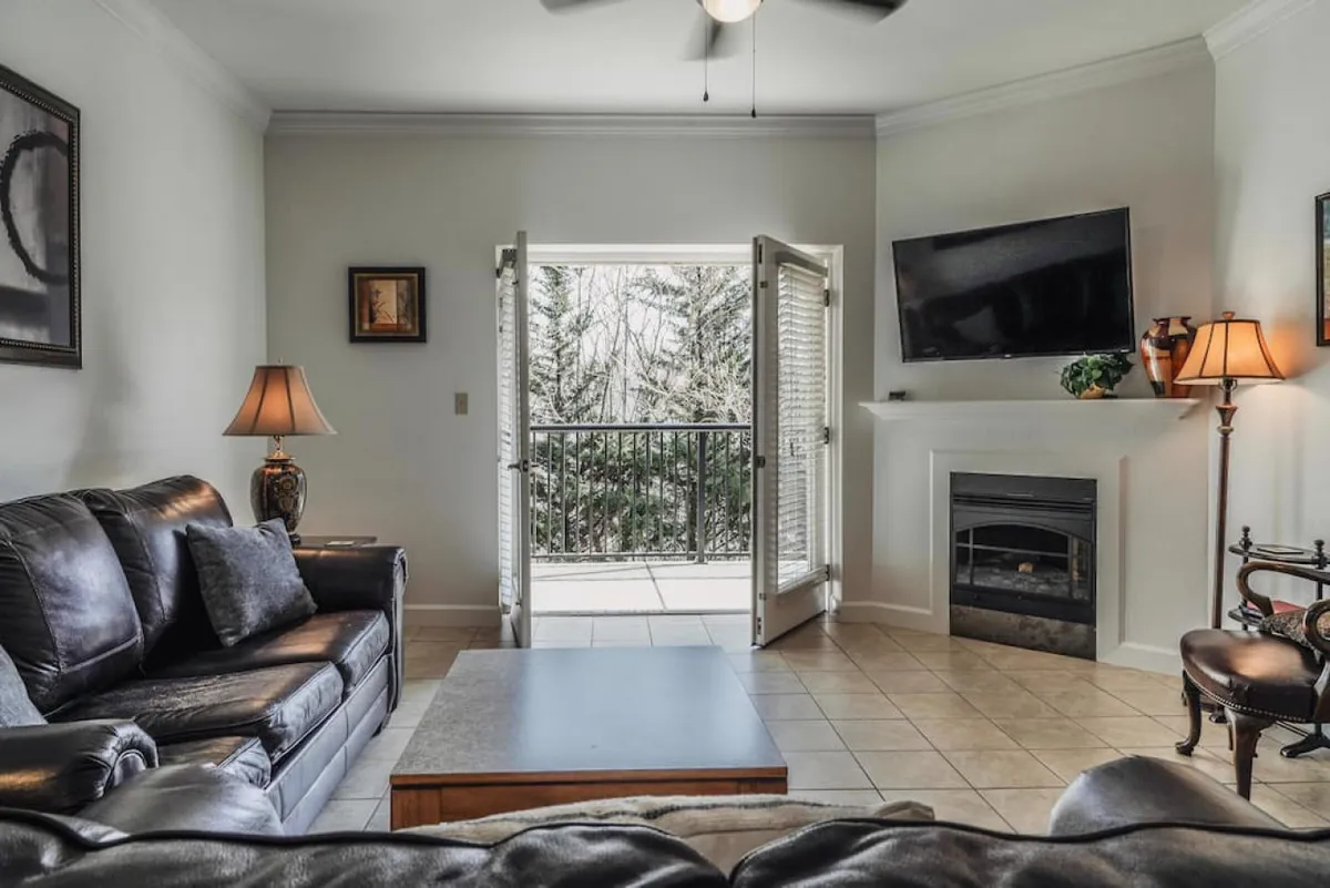 A spacious living room filled with natural sunlight streaming through open doors, a cozy sofa, coffee table, and a fireplace. The open doors visually connect the indoor and outdoor spaces, reflecting the fusion of privacy, relaxation, and convenience.