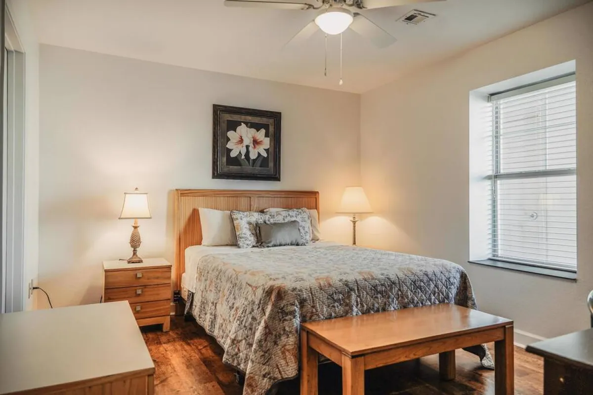 The image shows a serene bedroom with a cozy bed dressed in a patterned quilt, a soft-glowing lamp atop a nightstand, framed floral artwork adding a touch of charm, and a large window with blinds letting in gentle morning light.