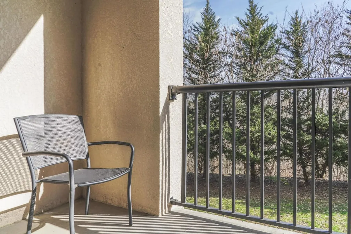 A serene balcony scene featuring a black metal chair, sturdy railing, and green trees in the background. The environment encourages relaxation, reading, or sipping a beverage while appreciating the view and tranquility.