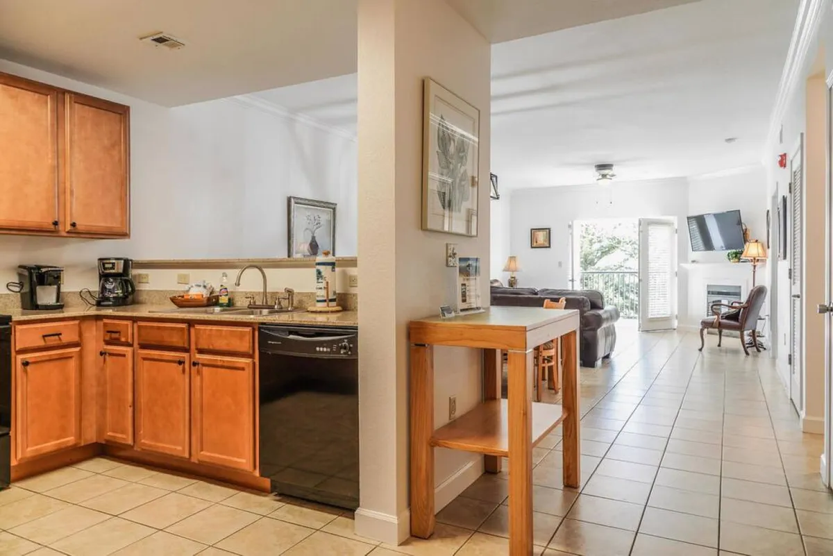 An inviting open-concept space with kitchen cabinetry, a countertop, and living room seating. Balcony doors in the background let in natural light and hint at mountain views, while the area is arranged for seamless entertaining, family meals, and game nights.