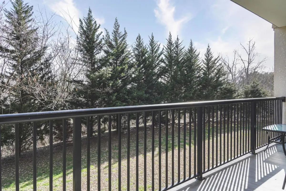 A view from a balcony showing a railing, dense trees, and an open sky, evoking a peaceful and private outdoor space. The setting suggests enjoying fresh air, relaxing with a morning coffee, and feeling connected to the surrounding environment.