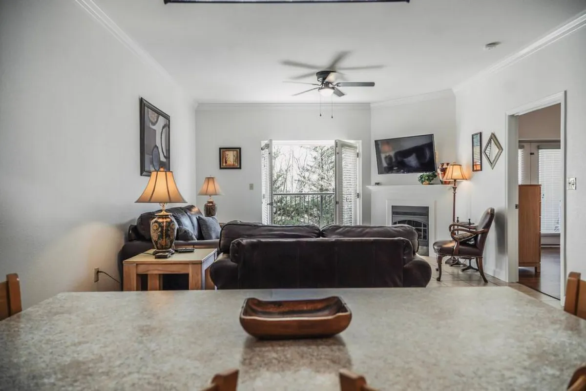 A spacious living room anchored by a plush sofa and coffee table, facing a large flat screen TV. Soft lighting from lamps and ample sunlight from a window create a welcoming atmosphere, while the arrangement encourages both relaxing and gathering for activities like movie nights.