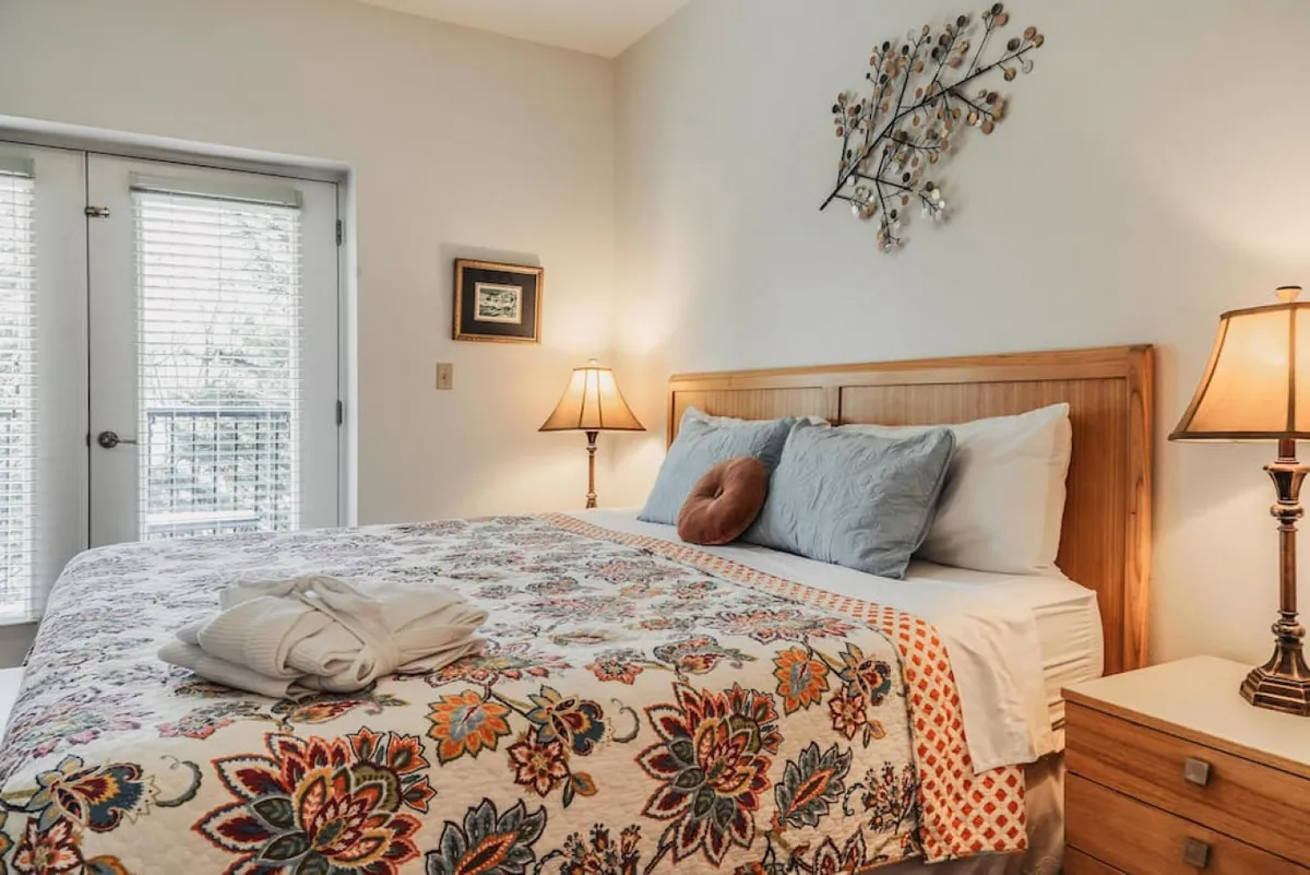 A cozy bedroom with patterned bedding, warm lighting, and a balcony door letting in natural light. The scene suggests a peaceful retreat where guests can enjoy relaxing mornings or evenings, with views extending outdoors to the changing landscape beyond.