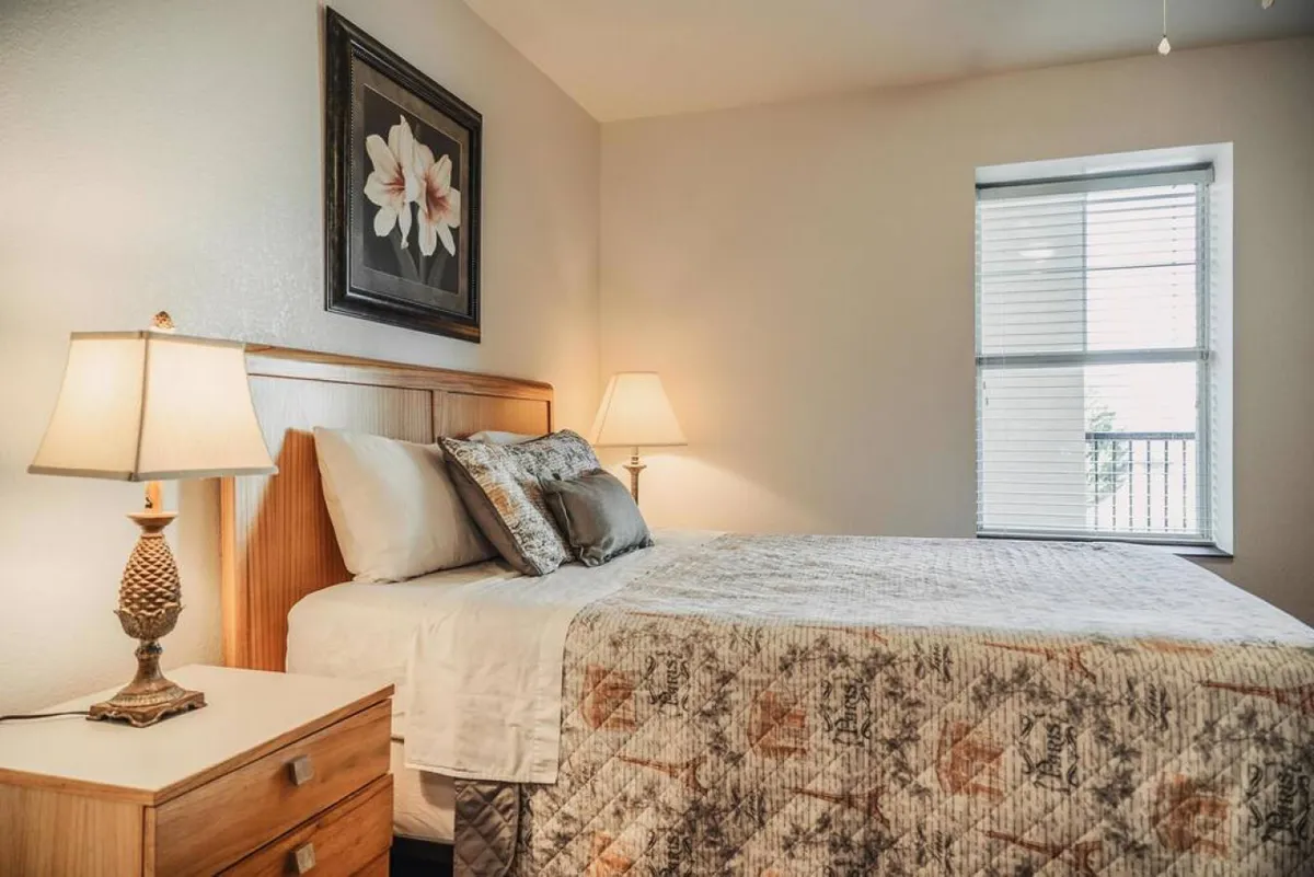 A serene bedroom with a large bed adorned with decorative pillows and a cozy quilt, flanked by nightstands with matching lamps. Framed artwork and a window with blinds complete the calm, homely setting, ideal for relaxing or reading.